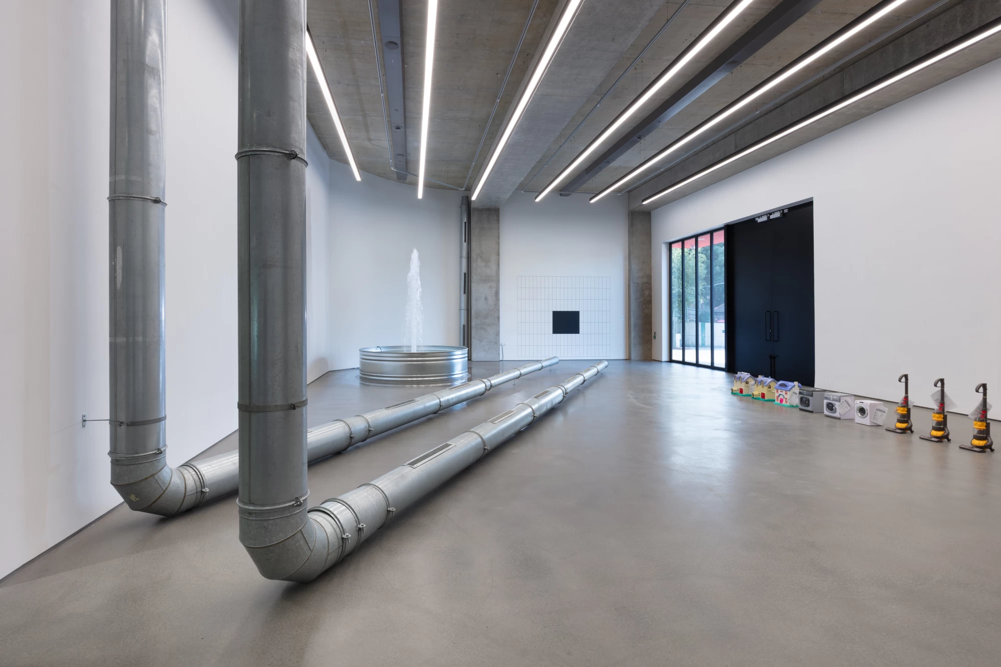 Large parts of metal tubing from an old air conditioning unit are assembled in two parallel 'L' shapes from the ceiling and across the floor. There is a water fountain with a metal base in the background, and a neat row of children's toys. The toys are all replicas of household items, like washing machines or vacuum cleaners. 